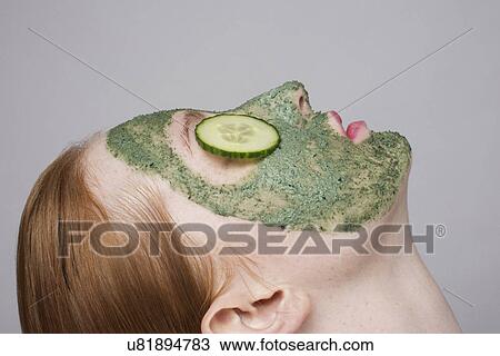 Stock Image - Young woman wearing face mask and cucumber over eyes. Fotosearch