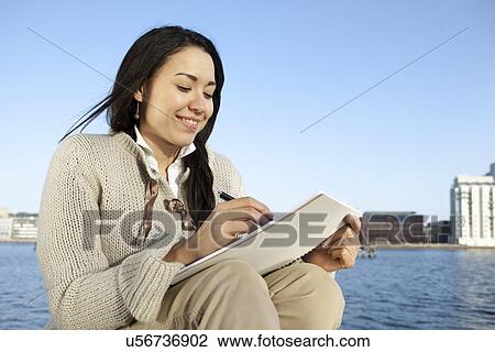 Stock Image - Young woman writing notes by waterfront, Copenhagen, Denmark. Fotosearch