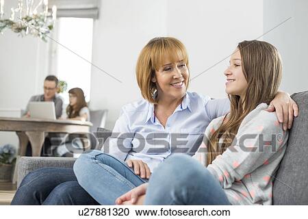 Stock Image - affectionate mother and daughter sitting on sofa with family in background. Fotosearch