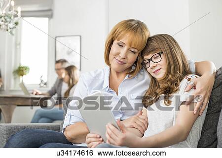 Affectionate mother and daughter using digital tablet with family sitting in background at home View Large Photo Image Stock Photograph - Affectionate mother and daughter using digital tablet with family sitting in background at home. Fotosearch