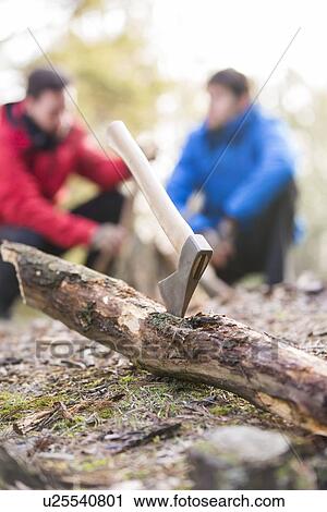 Stock Image - Axe stuck in log with male hikers in background at forest. Fotosearch
