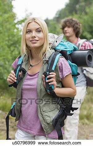 Stock Photo - Beautiful female backpacker looking away with man standing in background at forest. Fotosearch