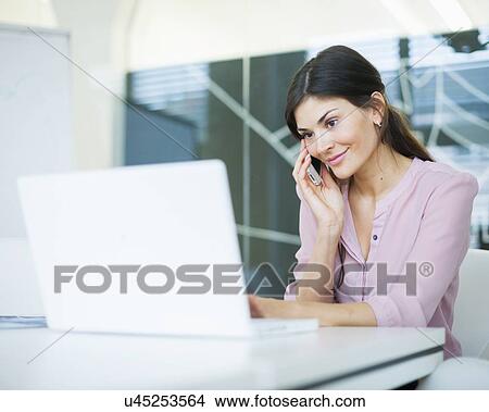 Picture - Beautiful young businesswoman using mobile phone while looking at laptop in office. Fotosearch