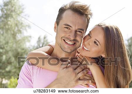 Stock Photo - Beautiful young woman embracing man from behind in park. Fotosearch