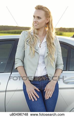 Stock Photo - Beautiful young woman looking away while standing by car at countryside. Fotosearch