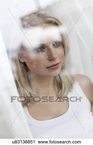 Stock Image - Beautiful young woman looking through hotel window. Fotosearch
