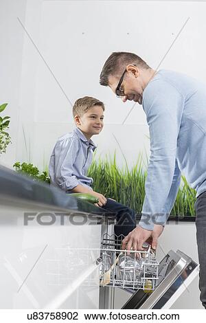 Stock Image - Boy looking at father placing glass in dishwasher at kitchen. Fotosearch
