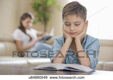 Boy reading book with mother in background at home View Large Photo Image Stock Photo - Boy reading book with mother in background at home. Fotosearch