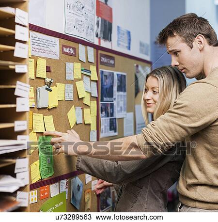 Stock Image - Business people reading reminders on bulletin board in office. Fotosearch