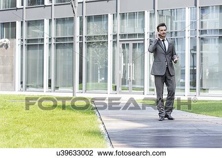 Businessman using cell phone while walking on path outside office View Large Photo Image Stock Image - Businessman using cell phone while walking on path outside office. Fotosearch