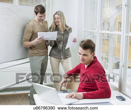 Stock Image - Businessman using laptop while coworkers discussing over documents in office. Fotosearch