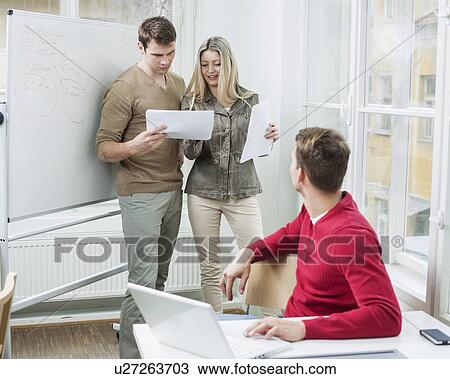 Stock Image - Businessman with laptop looking at coworkers discussing over documents in office. Fotosearch