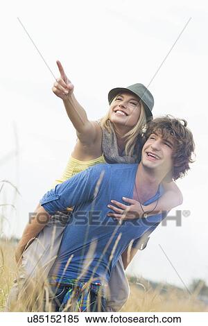 Stock Photography - Cheerful woman showing something while enjoying piggyback ride on man in field. Fotosearch