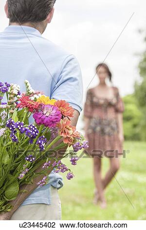 Stock Image - Cropped image of man surprising woman with bouquet in park. Fotosearch