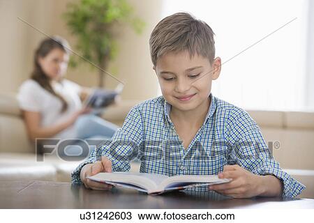 Cute boy reading book with mother in background at home View Large Photo Image Stock Image - Cute boy reading book with mother in background at home. Fotosearch