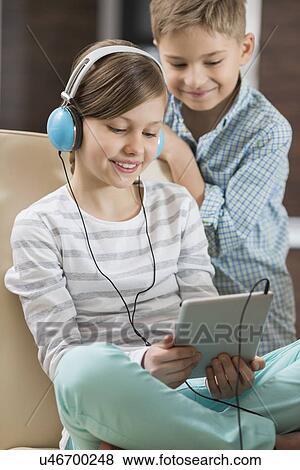 Cute girl listening music on digital tablet while brother standing behind her at home View Large Photo Image Stock Photo - Cute girl listening music on digital tablet while brother standing behind her at home. Fotosearch