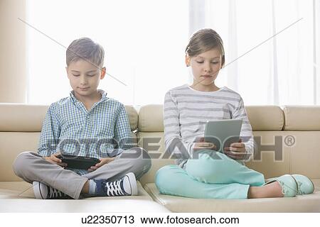 Stock Image - Cute siblings using technologies on sofa at home. Fotosearch