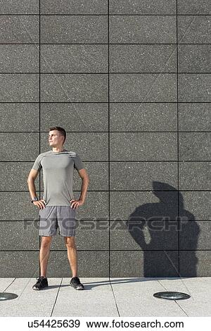 Stock Photo - Determined jogger standing with hands on hips against tiled wall outdoors. Fotosearch