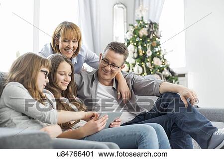 Stock Image - Family using tablet PC on sofa with Christmas tree in background. Fotosearch