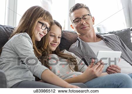 Stock Image - Father and daughters using tablet PC on sofa at home. Fotosearch