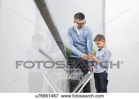 Stock Photo - Father and son placing glass in dishwasher at kitchen. Fotosearch