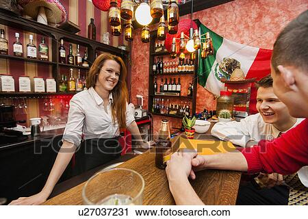 Stock Image - Female bartender with male customers at bar counter. Fotosearch