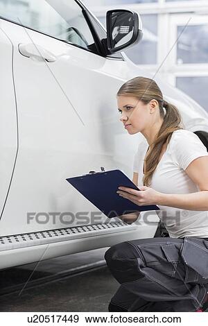 Stock Photo - Female mechanic holding clipboard while examining car body at repair shop. Fotosearch