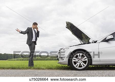 Picture - Frustrated young businessman using cell phone by broken-down car at countryside. Fotosearch
