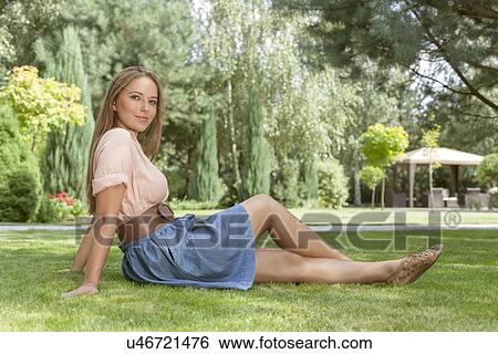 Stock Photograph - Full length of beautiful young woman looking away while relaxing on grass in park. Fotosearch