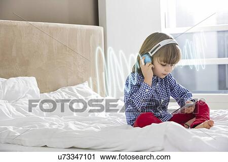 Stock Image - Full length of boy listening music through headphones on bed. Fotosearch