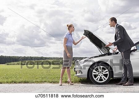 Stock Photo - Full length of business couple arguing by broken car at countryside. Fotosearch