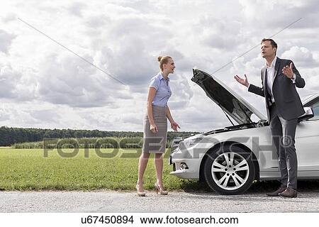 Picture - Full length of business couple having argument by broken car at countryside. Fotosearch