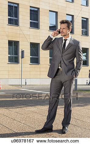 Stock Photo - Full length of businessman conversing on cell phone against office building. Fotosearch