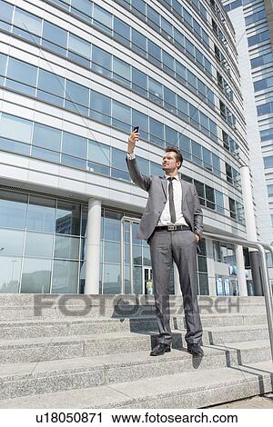 Stock Image - Full length of businessman taking selfie on steps outside office. Fotosearch