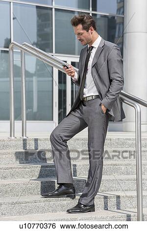 Full length of businessman text messaging through cell phone while standing on steps outside office View Large Photo Image Stock Photograph - Full length of businessman text messaging through cell phone while standing on steps outside office. Fotosearch
