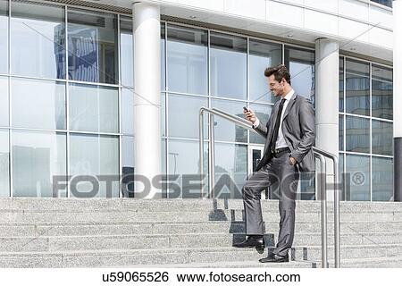 Stock Photograph - Full length of businessman text messaging through cell phone while standing on steps outside office. Fotosearch