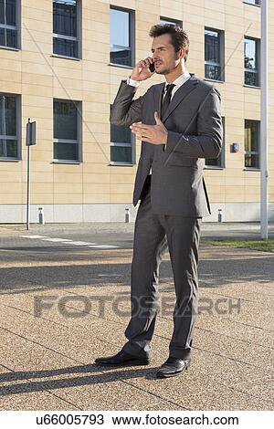 Stock Image - Full length of businessman using cell phone against office building. Fotosearch