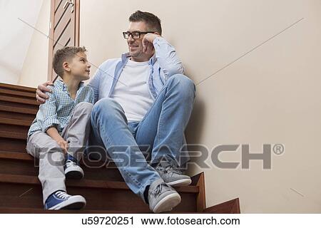 Stock Image - Full length of father and son looking at each other while sitting on steps at home. Fotosearch