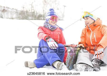 Stock Photograph - Full length of female friends with snowboard relaxing during winter. Fotosearch
