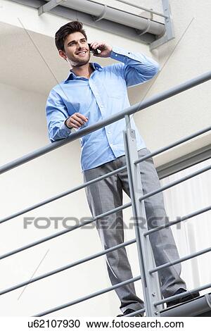 Stock Image - Full length of happy young businessman using mobile phone at balcony. Fotosearch