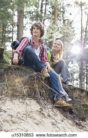 Stock Image - Full length of hiking couple sitting on edge of cliff in forest. Fotosearch
