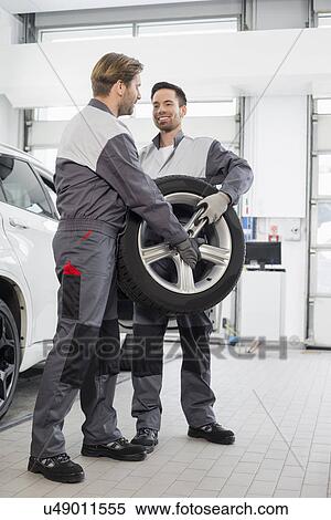 Stock Photography - Full length of maintenance engineers carrying tire in car workshop. Fotosearch