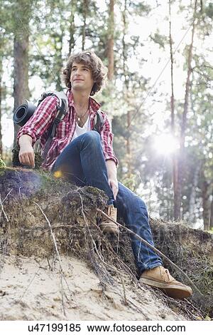 Stock Photography - Full length of male hiker looking away while sitting on cliff in forest. Fotosearch
