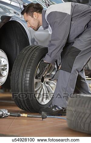 Stock Image - Full length of male mechanic fixing car's tire in repair shop. Fotosearch