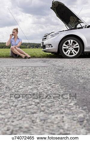 Stock Photography - Full length of mid adult businesswoman using cell phone while sitting beside broken down car at countryside. Fotosearch