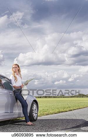 Stock Photography - Full length of mid adult woman leaning on car while holding map at countryside. Fotosearch
