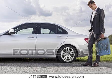 Stock Photo - Full length of sad young businessman carrying gas can by broken down car at countryside. Fotosearch