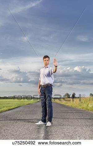 Full length of serious young man holding up hand to stop at country road View Large Photo Image Stock Image - Full length of serious young man holding up hand to stop at country road. Fotosearch