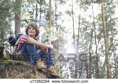 Full length of smiling male hiker looking away while sitting on cliff in forest View Large Photo Image Picture - Full length of smiling male hiker looking away while sitting on cliff in forest. Fotosearch