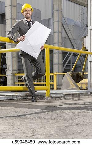 Full length of smiling young male architect holding blueprint outside industry View Large Photo Image Stock Photo - Full length of smiling young male architect holding blueprint outside industry. Fotosearch